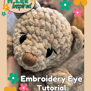 May include: Close-up of a crocheted stuffed animal's face with embroidered eyes. The animal is brown and has a brown nose. The text "Embroidery Eye Tutorial" is below the animal's face.