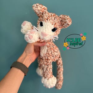 May include: A handmade crochet cat plushie in shades of cream, orange, and white. The cat has blue eyes, pink accents on the paws and nose, and white whiskers. The plushie is held up in front of a teal background. The text "H'Lee Inspired" is in the background.