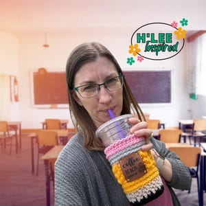 May include: A woman in a classroom setting holds a crocheted coffee cup cozy with the text "Coffee Teach Repeat" on a brown leather patch. The cozy is yellow, pink, gray, and black.