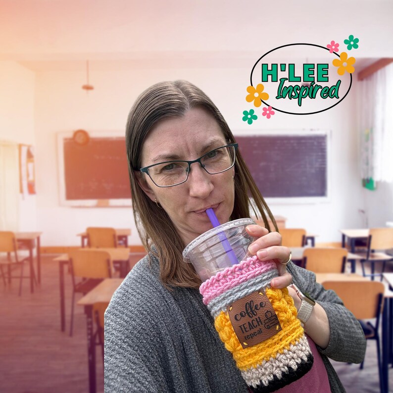 May include: A woman in a classroom setting holds a crocheted coffee cup cozy with the text "Coffee Teach Repeat" on a brown leather patch. The cozy is yellow, pink, gray, and black.