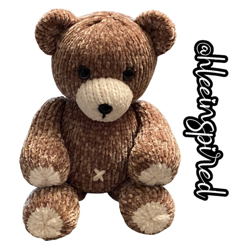 Op de afbeelding: Een bruine gebreide teddybeer met witte accenten op zijn poten en snuit. De beer zit met zijn poten voor zich. De beer is gemaakt van zacht, pluche materiaal.