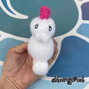 May include: White crocheted stuffed animal with a pink crocheted hat and black eyes. The animal is being held in a hand.