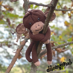 May include: A brown knitted monkey toy with a long tail, hanging from a tree branch. The monkey has a brown body, a light brown face, and a pink nose.
