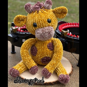 Pode incluir: Um brinquedo de girafa de crochê em amarelo e marrom. A girafa tem um nariz marrom e grandes olhos marrons. O brinquedo está sentado em uma superfície de madeira. @hleeinspired