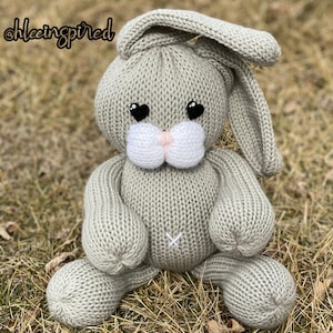 May include: A gray knitted bunny rabbit toy with black heart-shaped eyes and a white nose. The bunny is sitting on a bed of dry grass.