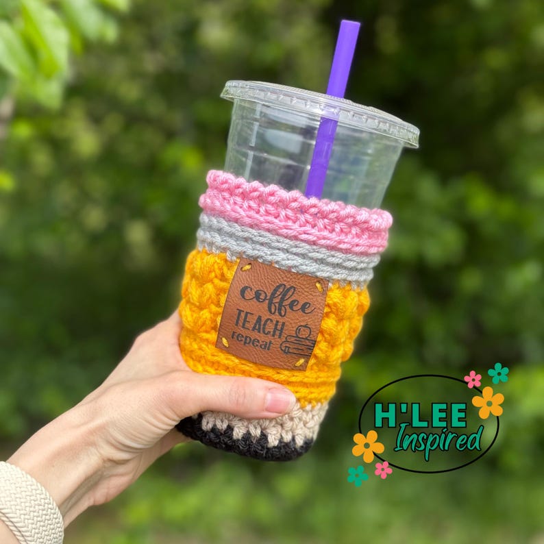 May include: A yellow, gray, pink, and black crocheted coffee cup cozy shaped like a pencil. The cozy has a brown leather patch with the text "coffee teach repeat" and a small apple graphic.