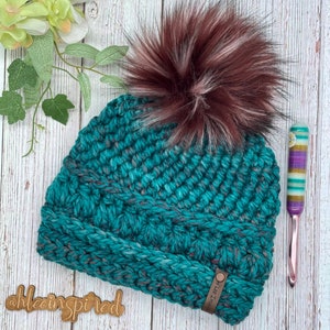 May include: Teal and burgundy crocheted beanie with a faux fur pom pom. The beanie has a textured stitch pattern and a leather tag that says "Hlee Inspired".