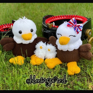Könnte beinhalten: Drei gestrickte Adler-Plüschtiere, eines mit einer rot-weiß-blauen Schleife. Die Adler sind braun und weiß mit gelben Füßen. Die Plüschtiere sitzen auf einem Bett aus grünem Gras.