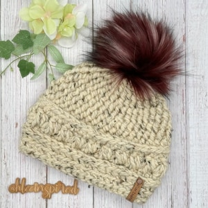 May include: A cream-colored crocheted beanie with a brown and white faux fur pom pom. The beanie has a cable knit pattern and a small leather tag that says "Ohlee Inspired".