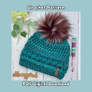 May include: Teal crochet hat with a brown and white faux fur pom pom. The hat has a textured stitch pattern and a brown leather tag with the text "#theinspired".