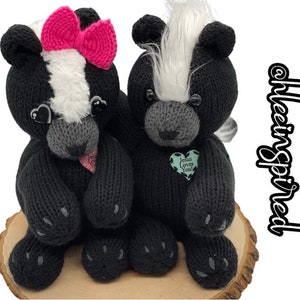 May include: Two knitted black and white skunk plushies sitting on a wooden slice. The skunk on the left has a pink bow and the skunk on the right has a green heart with the text "Jesus Loves You".