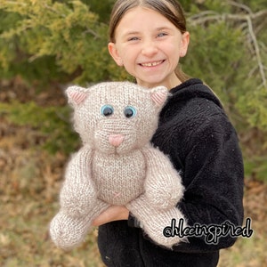 May include: A young person holding a knitted plush toy cat with pink ears, a pink nose, and blue eyes. The cat is light brown and white with a fluffy texture. The person is wearing a black hoodie with the text "@hleeinspired" on the bottom right corner.
