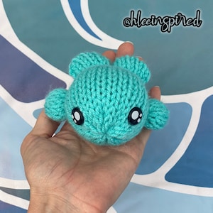 May include: A turquoise knitted creature with large, expressive eyes and small ear-like appendages. The handmade toy is held in a hand, showcasing its size and detail. The text "@khleeinspired" is in the upper right corner.