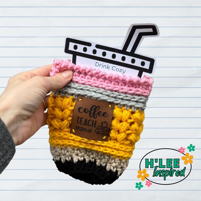 May include: A crocheted coffee cup cozy in yellow, gray, pink, and black with a brown leather patch that says "coffee teach repeat" and a black and white coffee cup graphic. The cozy is sitting on a white lined paper background with a white sticker that says "Drink Cozy" on top of the cozy.