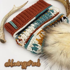 Può includere: Un cappello all'uncinetto con un motivo geometrico colorato e un pon pon in pelliccia sintetica. Il cappello ha un'etichetta in pelle marrone con il testo "Hilla Threads" su di essa. Il cappello è su una superficie di legno con un cartello di legno che dice "@hleeinspired".