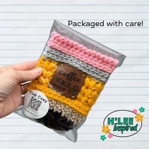 May include: A yellow, gray, and pink crocheted cup cozy shaped like a pencil. The cozy has a brown leather patch with the text "coffee teach repeat" and a coffee cup icon. The cozy is packaged in a clear plastic bag with a label that says "Cup Cozy" and has a QR code. The label also has the text "Scan to connect! h'leeinspired.com".