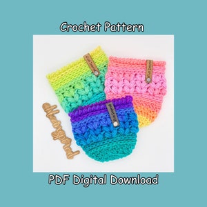 Könnte beinhalten: Drei gehäkelte Becherwärmer in Regenbogen-Ombre-Farben. Die Becherwärmer sind mit einem strukturierten Stich gearbeitet und haben jeweils ein braunes Leder-Etikett mit dem Text "Made Inspired".