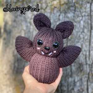 May include: A brown knitted bat with large green eyes and a white smile. The bat is held in a hand with the background out of focus.