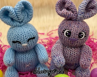 May include: Two knitted bunny plushies, one light blue and one purple with white speckles. Both have black eyes and a white nose. The bunnies are sitting on pink and white shredded paper.