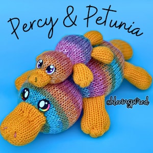Könnte beinhalten: Zwei gestrickte Plüschtiere in Form von Schnabeltieren. Das größere Schnabeltier ist blau, gelb und orange mit einem langen, gelben Schnabel. Das kleinere Schnabeltier ist rosa, lila und gelb mit einem kürzeren, gelben Schnabel. Das kleinere Schnabeltier sitzt auf dem größeren. Der Text "Percy & Petunia" ist in Schwarz über den Spielzeugen geschrieben.