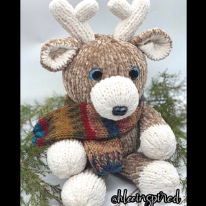 Könnte beinhalten: Ein gestricktes Rentier-Spielzeug mit braunem Fell, weißen Geweihen und einem bunten Schal. Das Rentier hat blaue Augen und eine schwarze Nase. Das Spielzeug sitzt auf einem Bett aus grünen Kiefernnadeln.