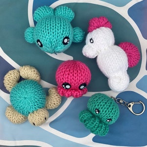 Peut inclure: Cinq jouets amigurumi en crochet en forme de créatures marines. Les jouets sont turquoise, blanc, rose et vert. Les jouets sont posés sur une surface à motifs colorés.