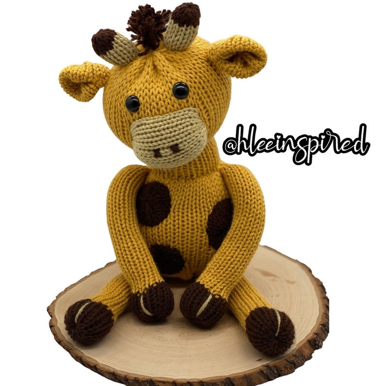 Op de afbeelding: Een gebreid geel giraffen speelgoed met bruine vlekken, zittend op een houten basis. De giraffe heeft een bruine neus, zwarte ogen en een bruine pluk haar op zijn kop. De tekst '@hleeinspired' is zichtbaar op de achtergrond.