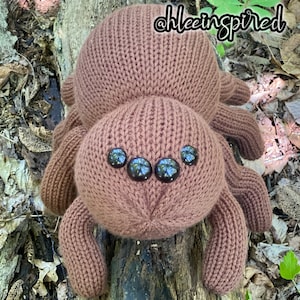 May include: A brown knitted spider with black button eyes. The spider is sitting on a tree trunk.