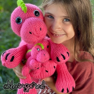 May include: A pink crocheted dinosaur with black claws and green accents. The dinosaur is holding a smaller pink crocheted dinosaur. A young person is holding the larger dinosaur.