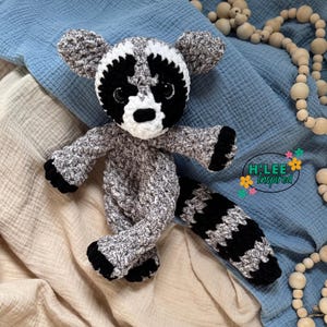 May include: A handmade crochet raccoon plush toy. The raccoon has a black and white striped tail, black paws, and a black mask around its eyes. The body is a speckled gray and white yarn. The text "H'Lee Inspired" is in the lower right corner.
