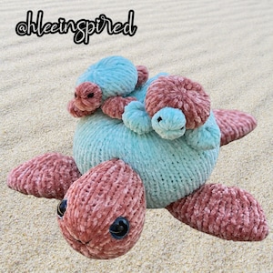 May include: A crocheted sea turtle toy with a light blue body and pink legs and head. Two smaller crocheted sea turtles, one light blue and one pink, are attached to the back of the larger turtle.