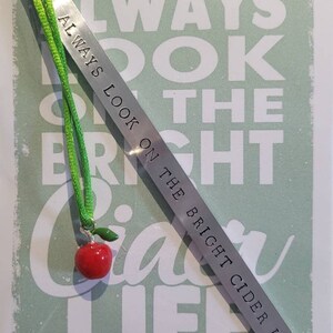 Könnte beinhalten: Silbernes Lesezeichen mit grünem Band und einem roten Apfel-Anhänger. Das Lesezeichen trägt die Gravur "Always look on the bright cider life!"