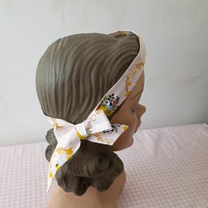Puede incluir: Una diadema blanca con un estampado floral y de abejas, atada con un lazo. La diadema está envuelta alrededor de una cabeza de maniquí. El diseño incluye flores y abejas amarillas y verdes.