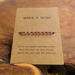 May include: A brown card with the text "MAKE A WISH" printed on it. A pink beaded bracelet is displayed on the card. The text below the bracelet reads "Put on this jewelry and make a wish. When the chain has worn through, legend says your wish will come true."