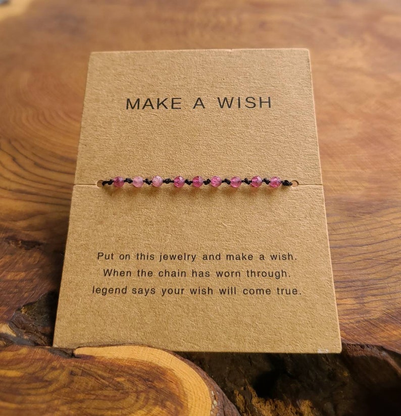 May include: A brown card with the text "MAKE A WISH" printed on it. A pink beaded bracelet is displayed on the card. The text below the bracelet reads "Put on this jewelry and make a wish. When the chain has worn through, legend says your wish will come true."