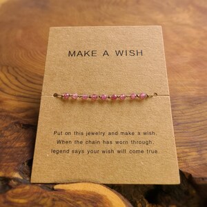 May include: A brown card with the text "MAKE A WISH" printed on it. A pink beaded bracelet is displayed on the card. The text below the bracelet reads "Put on this jewelry and make a wish. When the chain has worn through, legend says your wish will come true."
