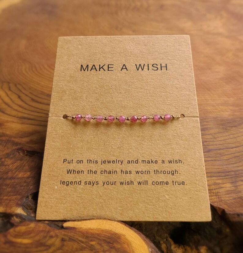 May include: A brown card with the text "MAKE A WISH" printed on it. A pink beaded bracelet is displayed on the card. The text below the bracelet reads "Put on this jewelry and make a wish. When the chain has worn through, legend says your wish will come true."