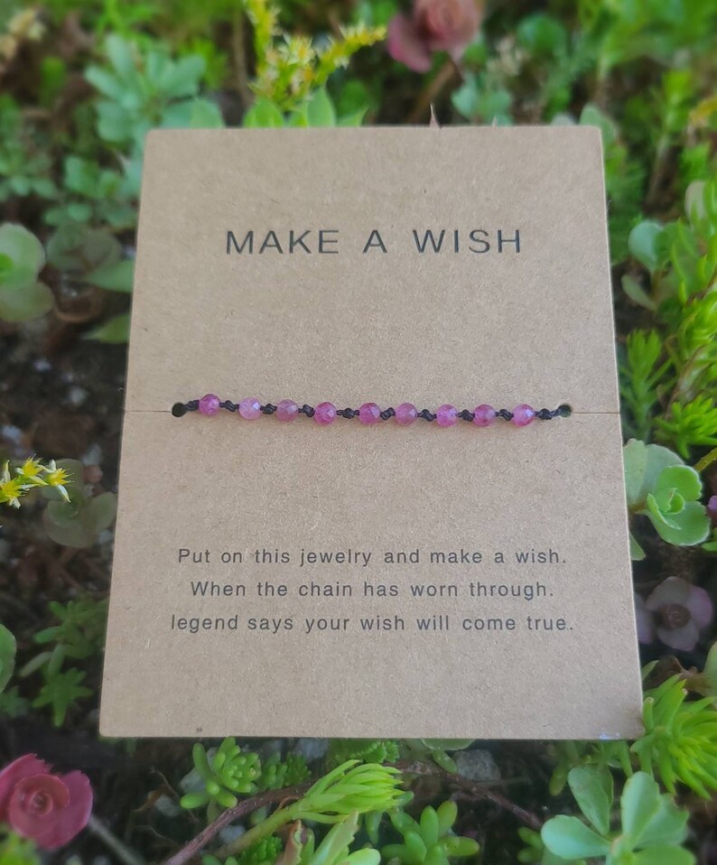 May include: A brown card with the text "MAKE A WISH" printed on it. A pink beaded bracelet is displayed on the card. The text below the bracelet reads "Put on this jewelry and make a wish. When the chain has worn through, legend says your wish will come true."