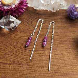 May include: A pair of silver dangle earrings with three small, round, purple beads on each earring.