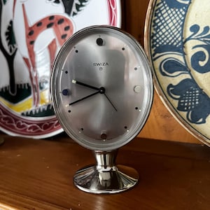 May include: A silver-coloured SWIZA 8 clock with an oval face and a pedestal base. The clock has black hands and a silver face with small black dots marking the hours. The clock is set on a wooden surface, with decorative plates in the background.