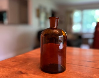 May include: A dark brown glass bottle with a wide mouth and a rounded bottom. The bottle is sitting on a wooden table.
