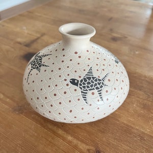 May include: A white ceramic vase with a round body and a narrow neck. The vase is decorated with a pattern of brown dots and black stylized birds.