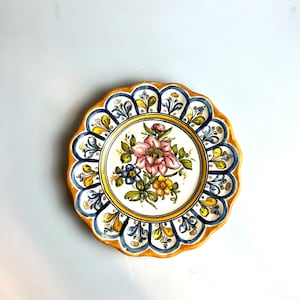 May include: A decorative ceramic plate with a scalloped edge and a floral design. The plate is painted in shades of blue, yellow, and pink.
