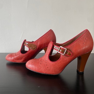 Peut inclure: Une paire de chaussures à talons Mary Jane en cuir rouge avec des détails marron. Les chaussures présentent une bride en T avec une boucle et une finition texturée. Les talons sont de hauteur moyenne et les chaussures ont des perforations décoratives.