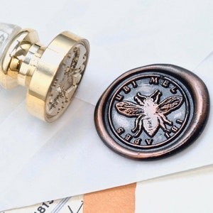 Könnte beinhalten: Ein Wachssiegelstempel mit einem goldfarbenen Metallstempelkopf und einem weißen und goldenen Griff. Der Stempelkopf zeigt ein Bienenmotiv mit den Worten "UBI MEL APES IBI" um die Biene herum. Ein dunkelbraunes Wachssiegel mit dem gleichen Bienenmotiv befindet sich neben dem Stempel.