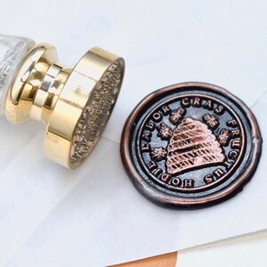 Könnte beinhalten: Ein goldfarbener Wachssiegelstempel mit einem durchsichtigen Glasgriff und einem bronzefarbenen Wachssiegel. Das Siegel zeigt ein Bienenkorb-Design mit den Worten "Labor Fructus" und "Hodie Cras" am Rand. Der Stempel liegt auf weißem Papier.