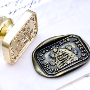 Könnte beinhalten: Ein goldfarbener Wachssiegelstempel und ein passendes Wachssiegel. Der Stempel hat einen transparenten Griff mit Goldflocken. Das Siegel ist dunkel mit goldfarbenen Buchstaben und einem Bienenkorbmotiv. Die Worte "HODIE LABOR CRAS FRUCTUS" sind sichtbar.