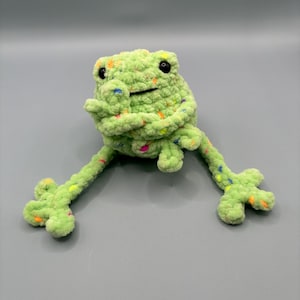 May include: A green crocheted frog with colorful speckles and black eyes. The frog is sitting with its legs outstretched.
