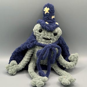 May include: A crocheted plush toy in the shape of a creature wearing a blue wizard hat with yellow stars and a blue cape. The creature has a gray body with black eyes and a black mouth.