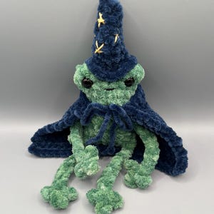 May include: A plush frog toy made of green textured yarn, wearing a dark blue wizard hat with yellow stitched star details and a matching dark blue cape tied at the neck. The frog has large black plastic eyes and long, dangling limbs.